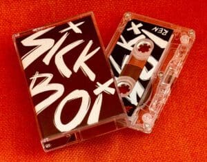 Cassette tape of Ren's 'Sick Boi' album with bold lettering on the cover, placed against a vibrant orange background.