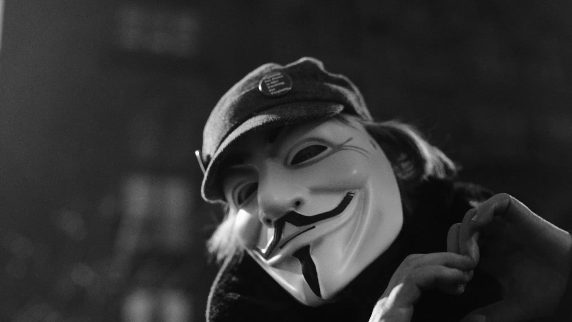 A close-up of an anonymous individual wearing a Guy Fawkes mask, forming a heart with their hands during the filming of RIOT’s “The Future Is Unwritten.”