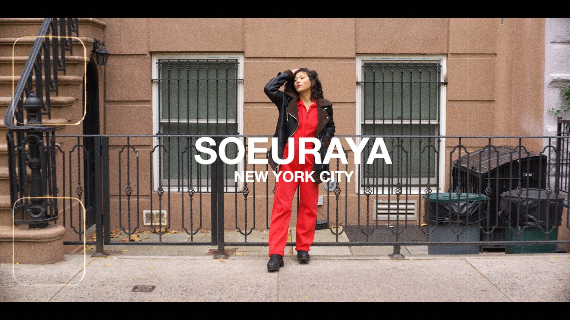 Soureya, poised and stylish in red jumpsuit and black jacket, stands confidently on a New York City street, embodying the urban energy and personal empowerment central to the SoulCycle Docufilm narrative