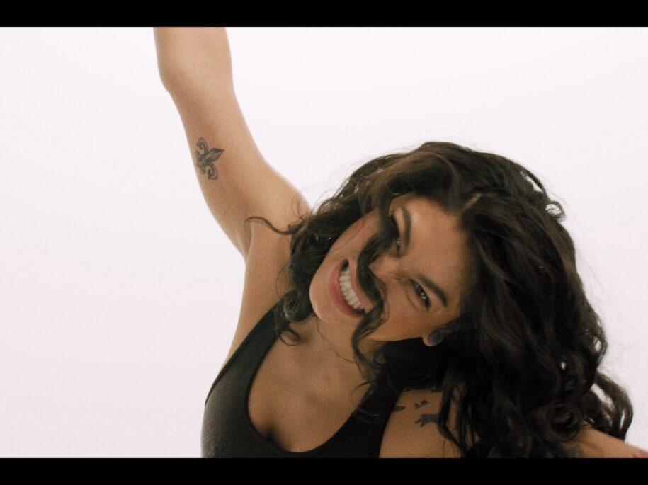 SoulCycle instructor Soureya celebrates with a joyous expression and raised arm, showcasing her tattoo, while wearing workout attire, representing the triumph and energetic spirit of SoulCycle's empowering workouts, as featured in the SoulCycle Docufilm