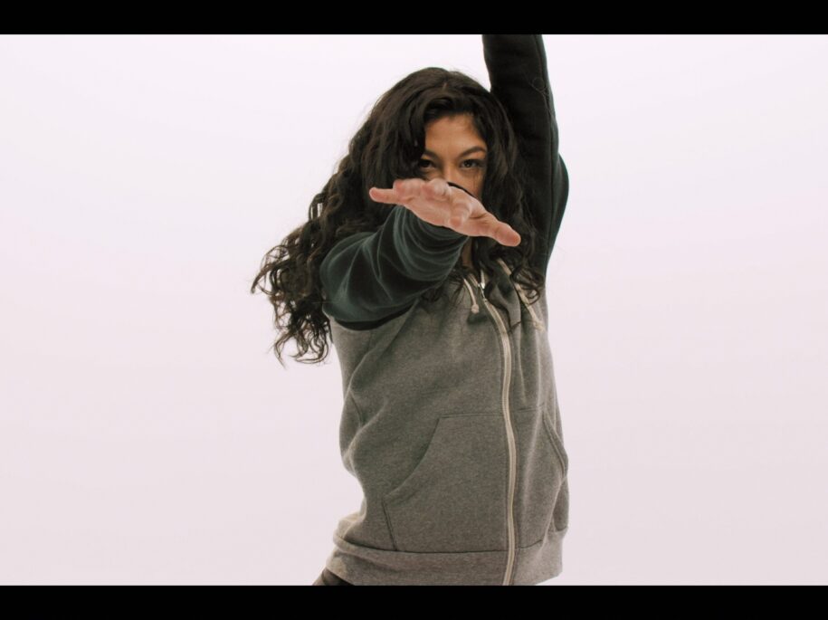 Soureya, SoulCycle instructor, strikes a dynamic dance pose in a casual gray hoodie, capturing the spirit of freedom and self-expression in a minimalist setting for the SoulCycle Docufilm, highlighting the personal joy and empowerment found in dance and fitness.