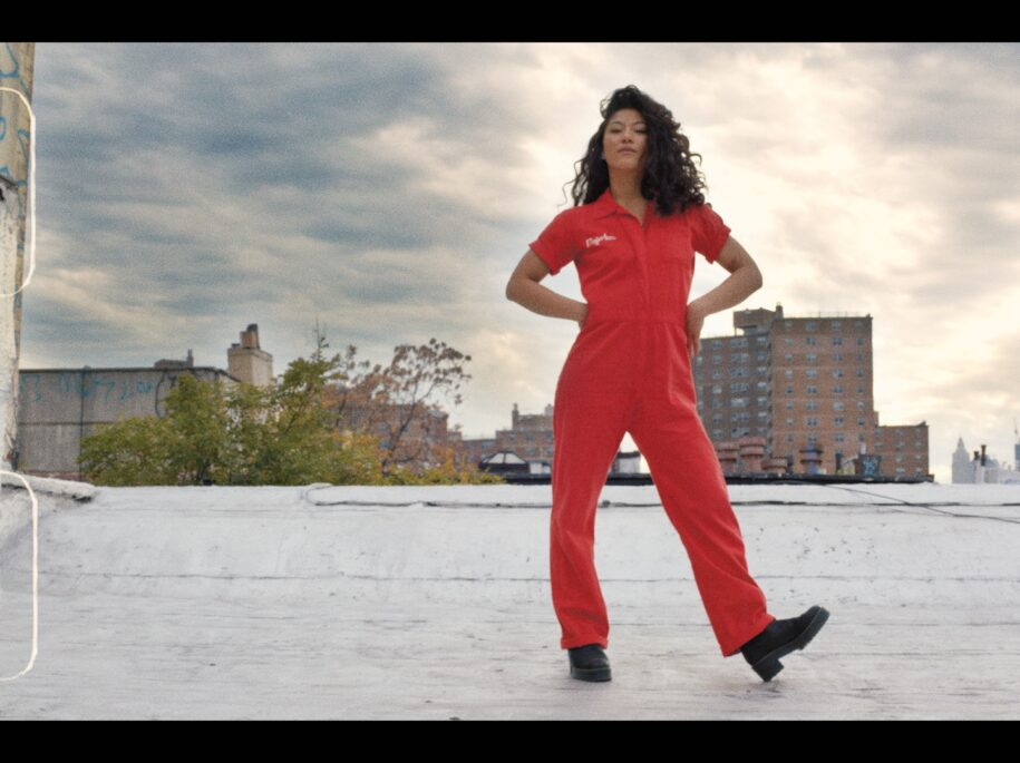 Soureya stands empowered on a rooftop in New York City, dressed in vibrant red, with the city skyline stretching into the distance, encapsulating the energetic and bold spirit of the SoulCycle Docufilm