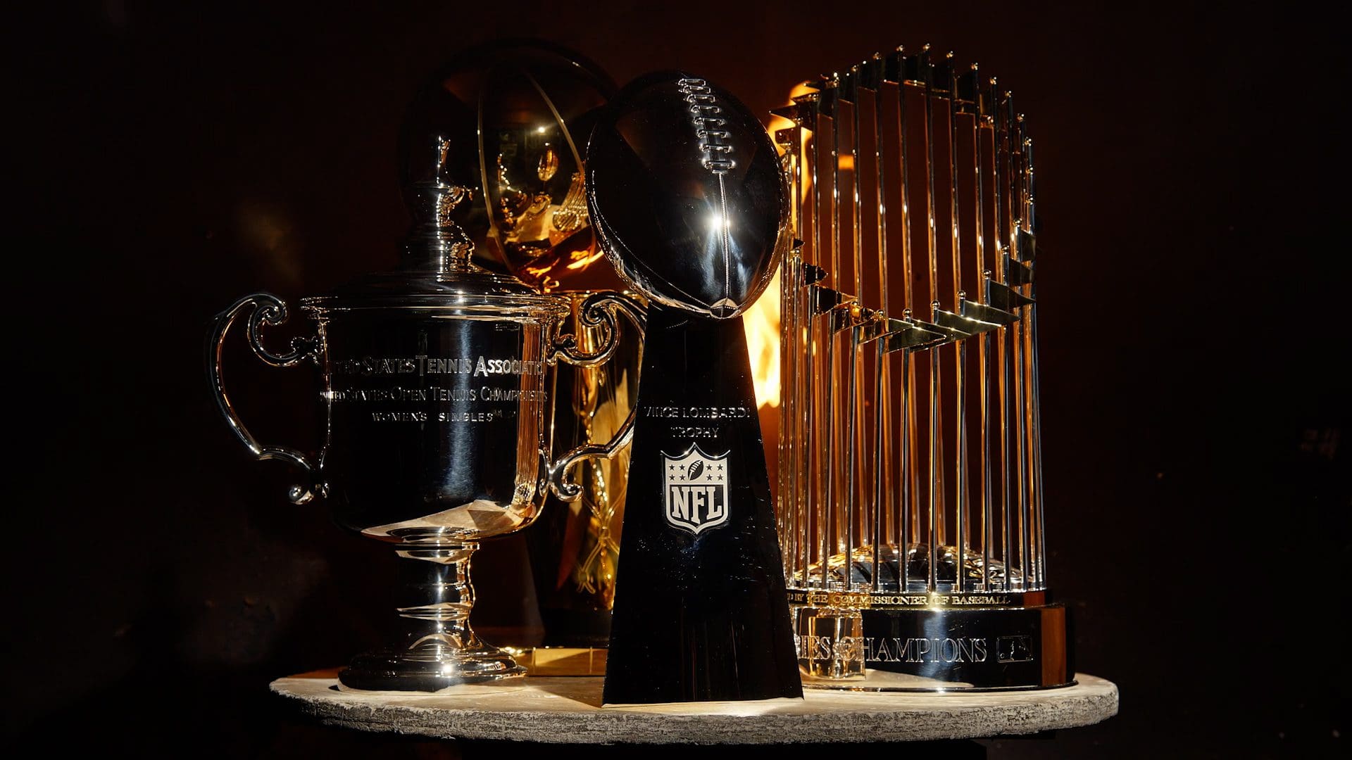 An array of prestigious sports trophies including the USTA Women's Singles trophy, the NFL's Vince Lombardi Trophy, and the MLB's Commissioner's Trophy, all crafted by Tiffany & Co., displayed against a dark background.