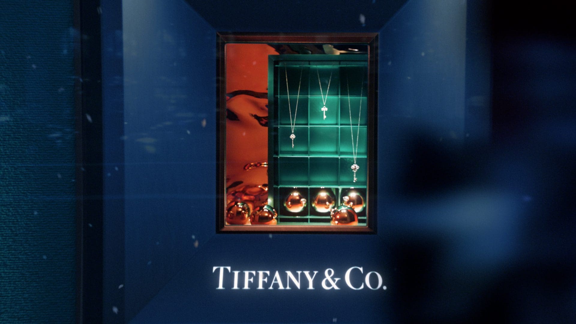 Elegant Tiffany & Co. window display featuring a selection of fine jewelry, including necklaces and pendants, set against a teal backdrop with the brand's signature in white text below.