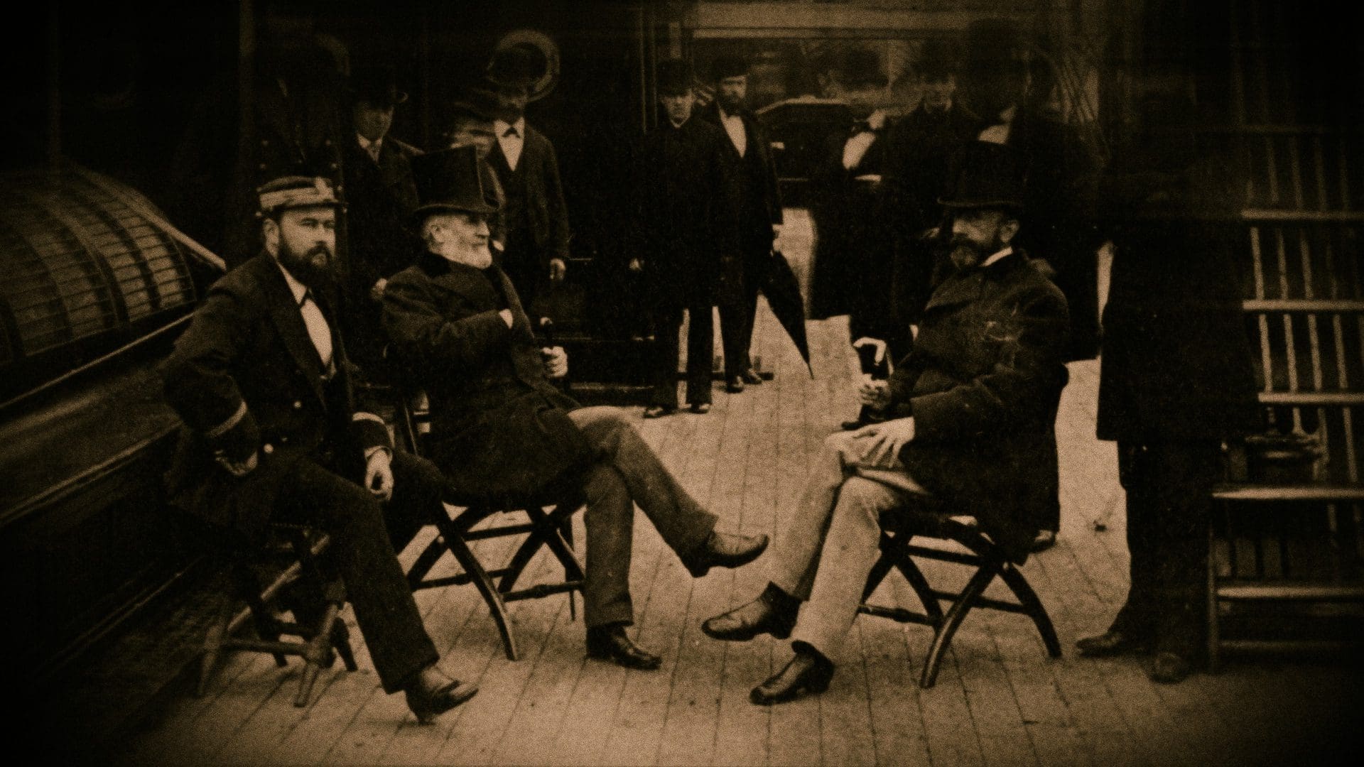 Sepia-toned historical photograph capturing Charles Lewis Tiffany and group of gentlemen in period attire, including top hats and suits, engaged in conversation while seated on folding chairs, likely depicting a scene from the late 19th or early 20th century.