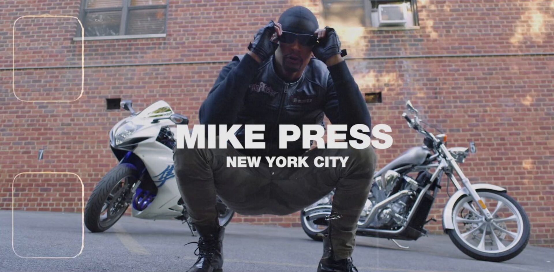 Mike Press crouching confidently in front of two motorcycles, wearing a leather jacket and sunglasses, set against an NYC brick wall.