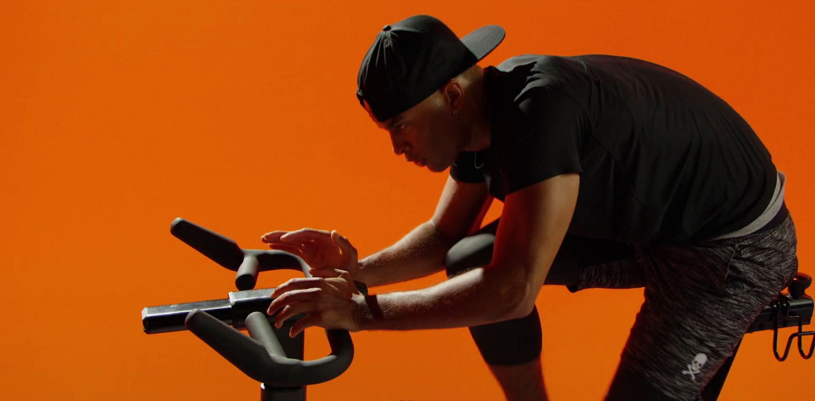 Mike Press riding a SoulCycle bike in a focused stance, set against a vibrant orange background.