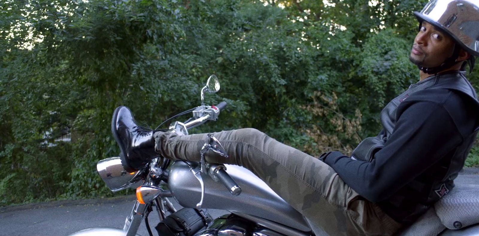 Mike Press relaxing on a motorcycle in camo pants and a reflective helmet, surrounded by greenery.